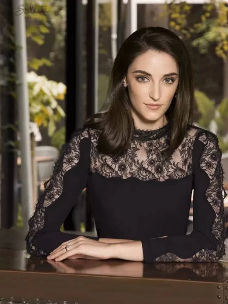 A model sitting at a table wearing a black long-sleeve top with a high-neck floral lace bodice and opaque sleeves.