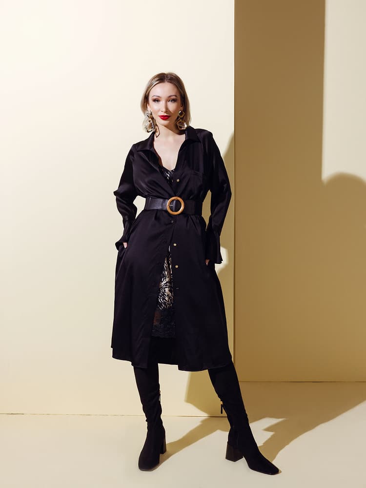 A model wearing a long black silk button-down maxi jacket cinched with a wide wooden buckle belt over a printed mini dress.