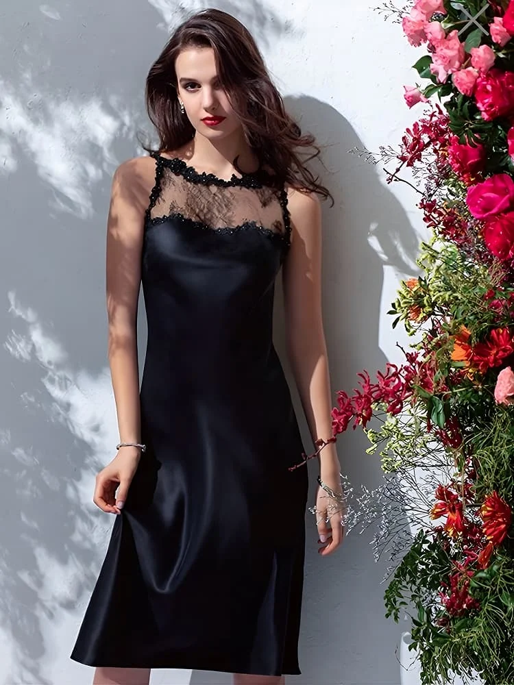 A model posing in a black silk slip dress featuring a sheer lace panel at the neckline adorned with black floral appliques.