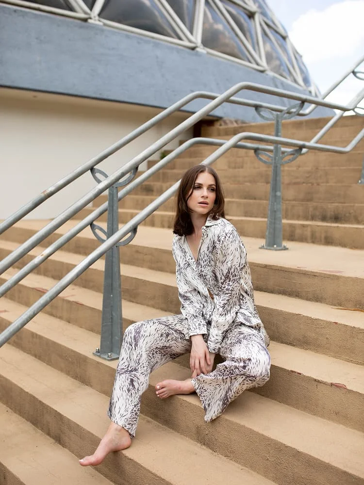 A model sitting on outdoor concrete steps wearing a contemporary grey and white abstract print silk pajama set.