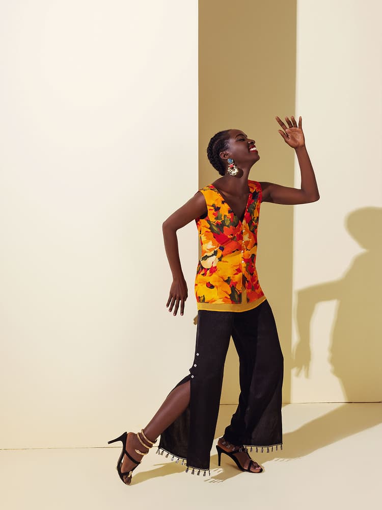 A model in a joyful pose wearing a sleeveless silk top with a large summer flower print, paired with black wide-leg trousers featuring tassel details.