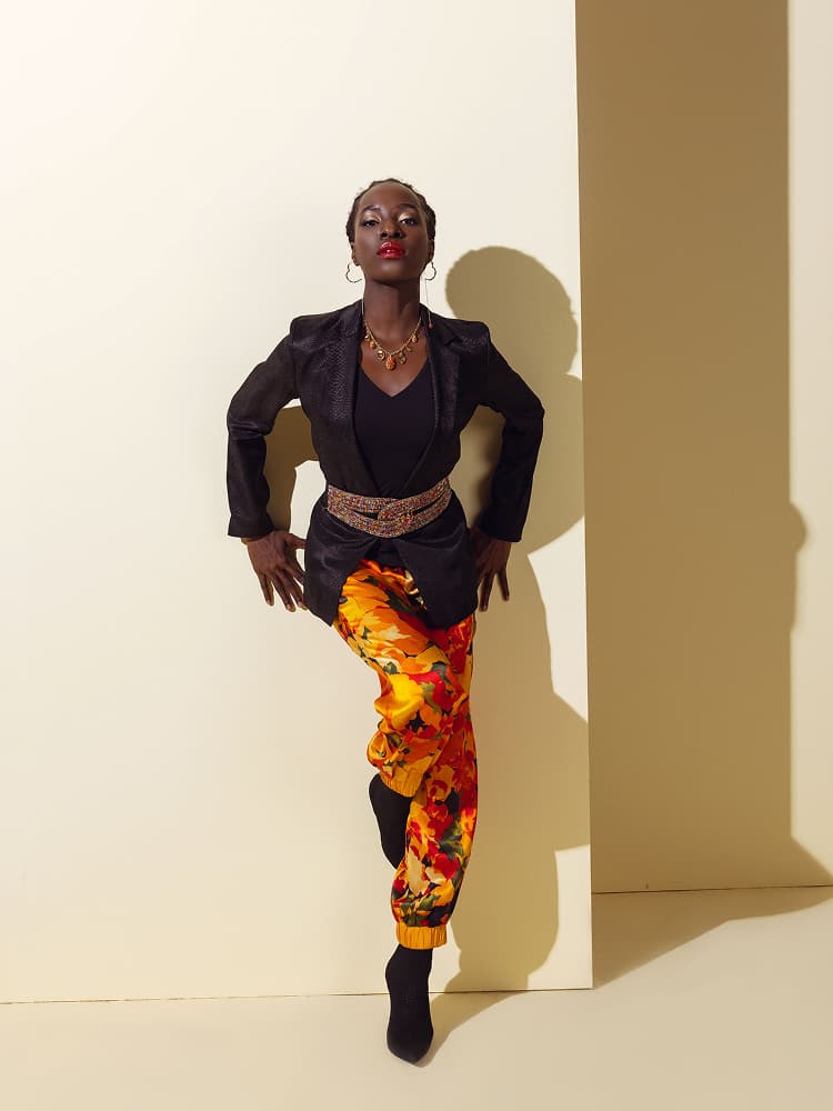 A model posing in vibrant yellow and orange summer flower print silk joggers, cinched with a jeweled belt and topped with a black tailored jacket.