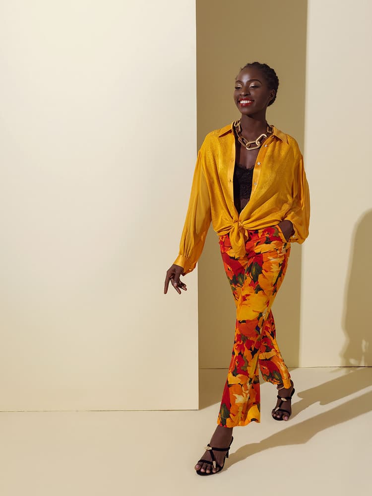 A model walking in a sheer yellow silk button-down shirt tied at the waist, paired with matching summer flower print silk trousers.